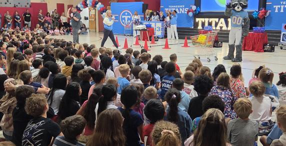 A large crowd of children watches an event featuring a mascot and a speaker.