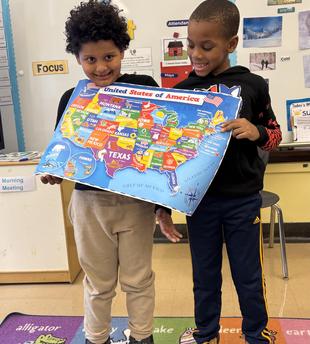 Two children holding a colorful map of the United States in a classroom.