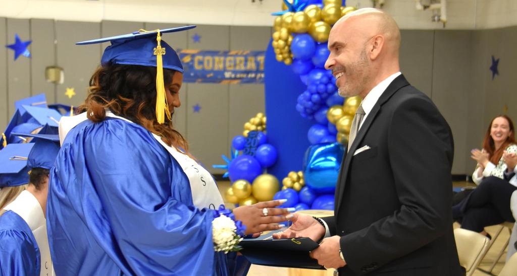 A graduate in blue gown receiving a diploma with a smiling man in a suit.