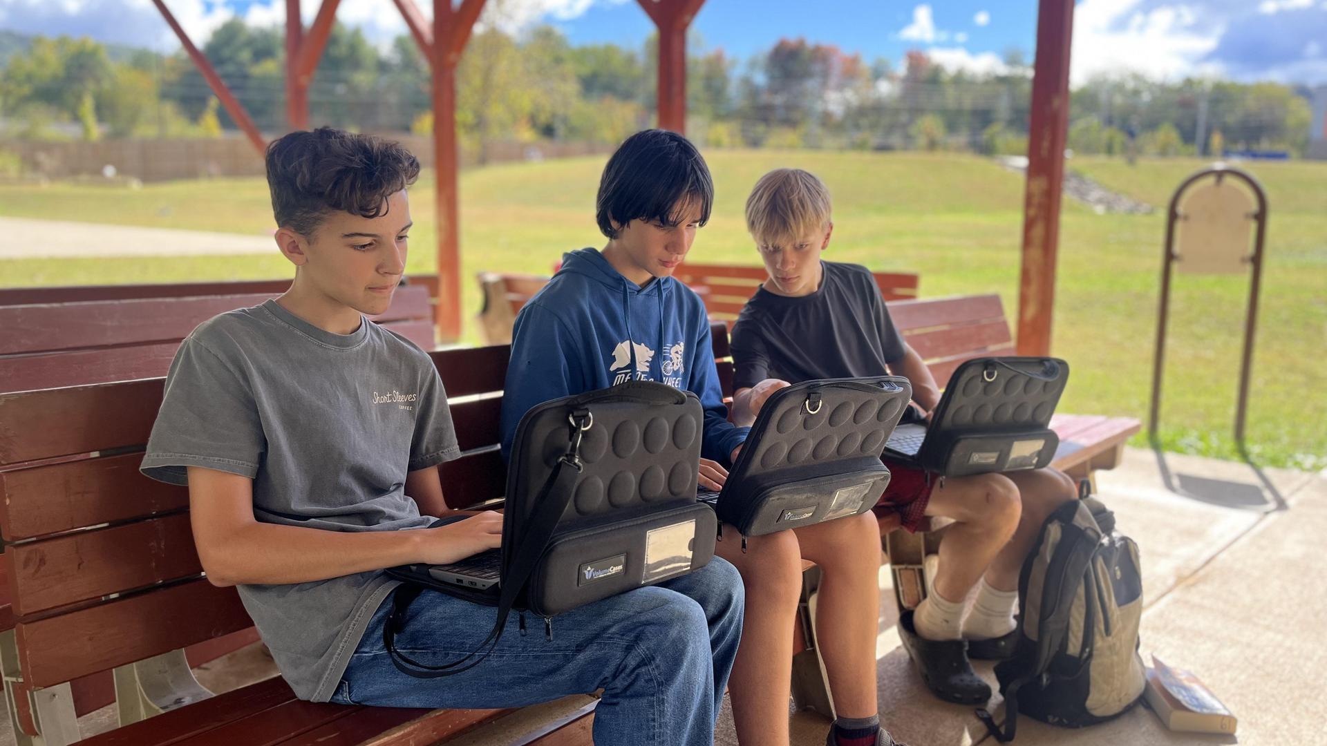 Three boys sitting on a bench outdoors, working on computers