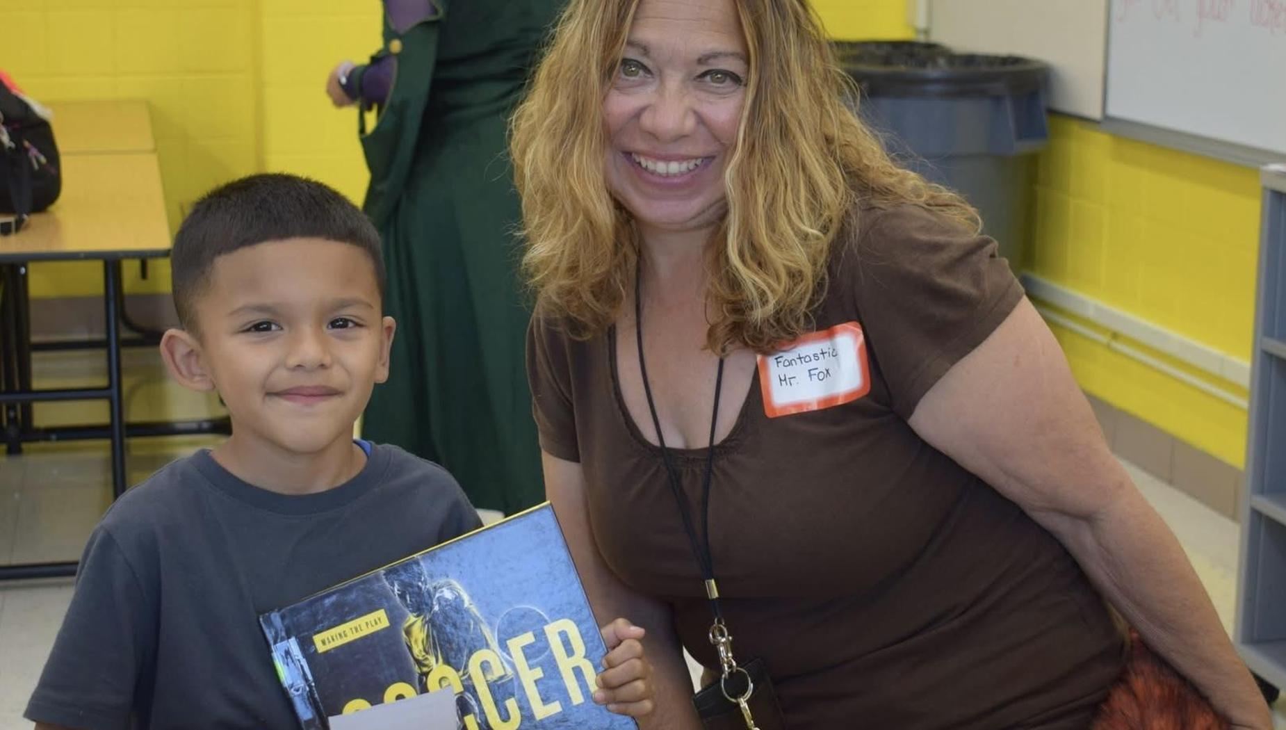 A boy holding a soccer book poses with a smiling teacher in a classroom.