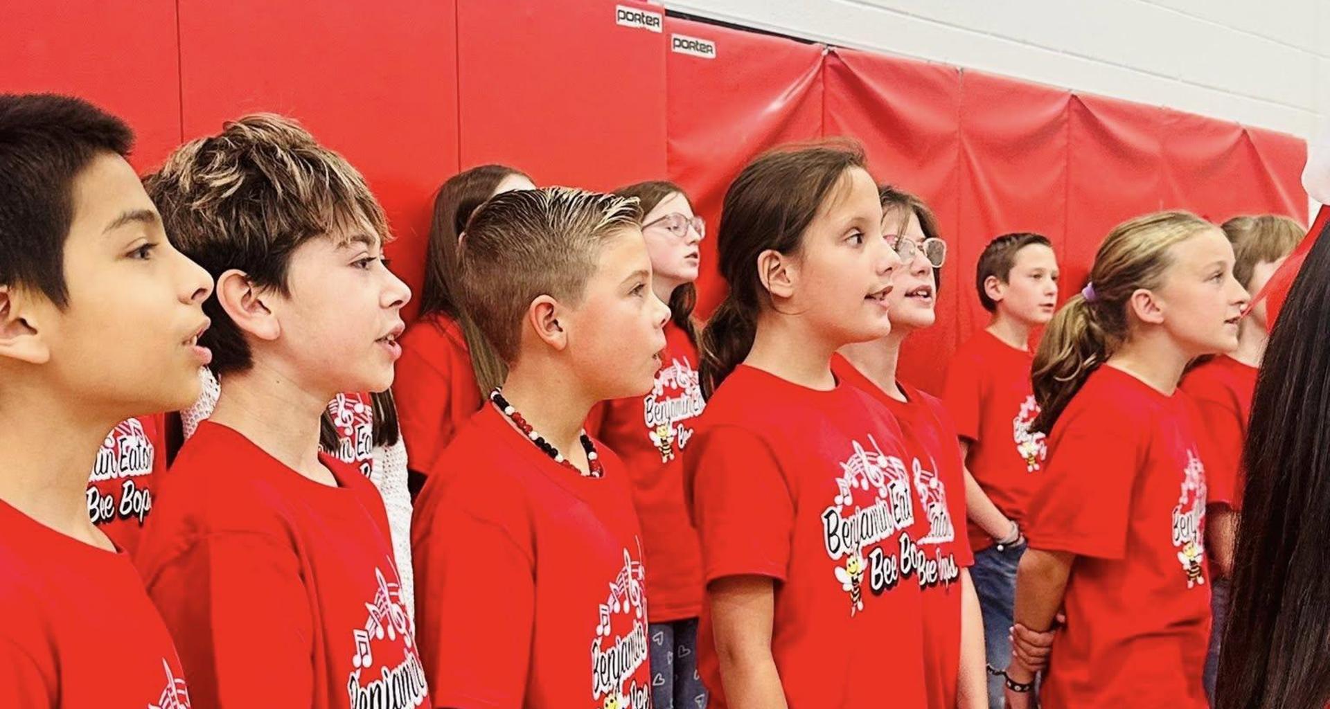 Children in matching red t-shirts singing together in a gymnasium setting.