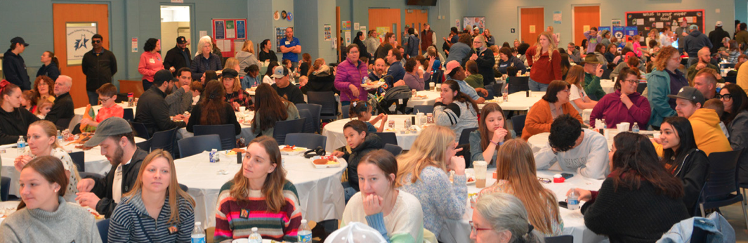 A large gathering of people enjoying the Texas School for the Deaf (TSD) annual Pancake Breakfast. Families, students, and community members sit at round tables covered with white tablecloths, chatting and eating. The background shows a bustling atmosphere with more attendees lining up and mingling near the walls, decorated with posters and artwork. The event exudes a warm, community-centered vibe.