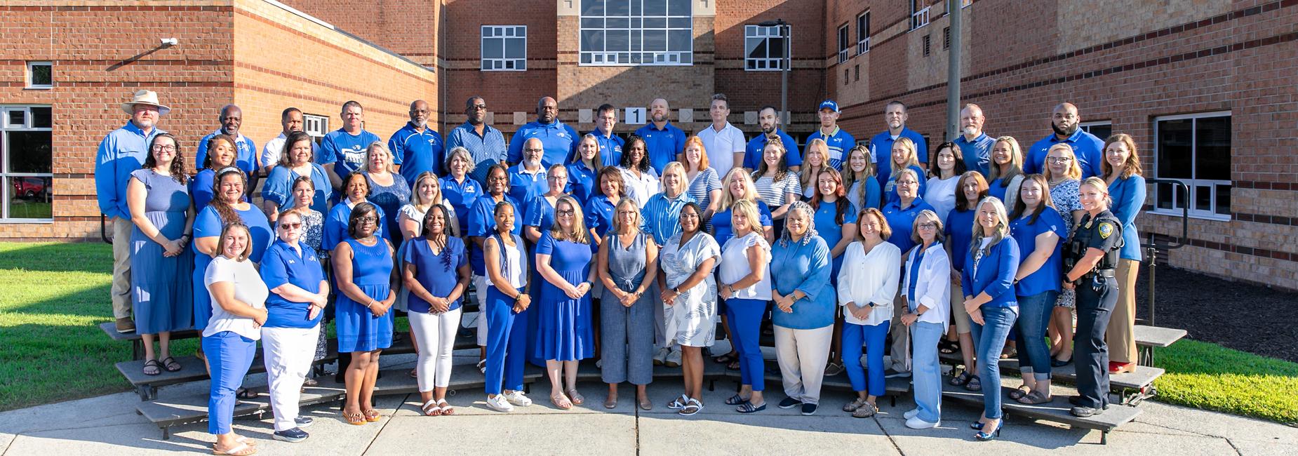 Group photo of a large diverse staff in blue attire outside a school building.