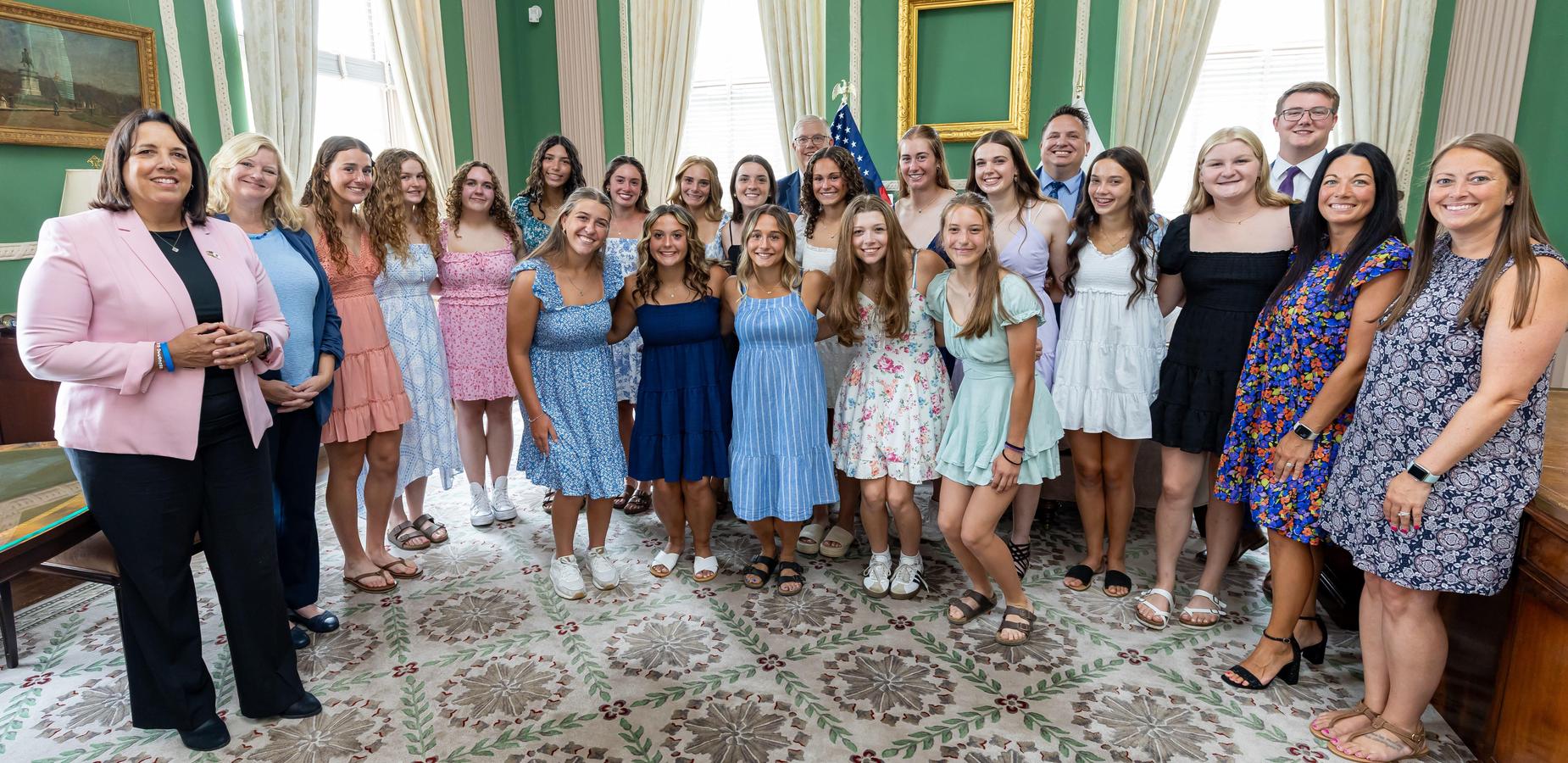A group of smiling teenage girls and their coaches stand together in a formal room with green walls, large windows, and a patterned rug. The girls are dressed in a variety of colorful dresses, while the adults are in a mix of professional and casual attire. A few men are visible in the background.