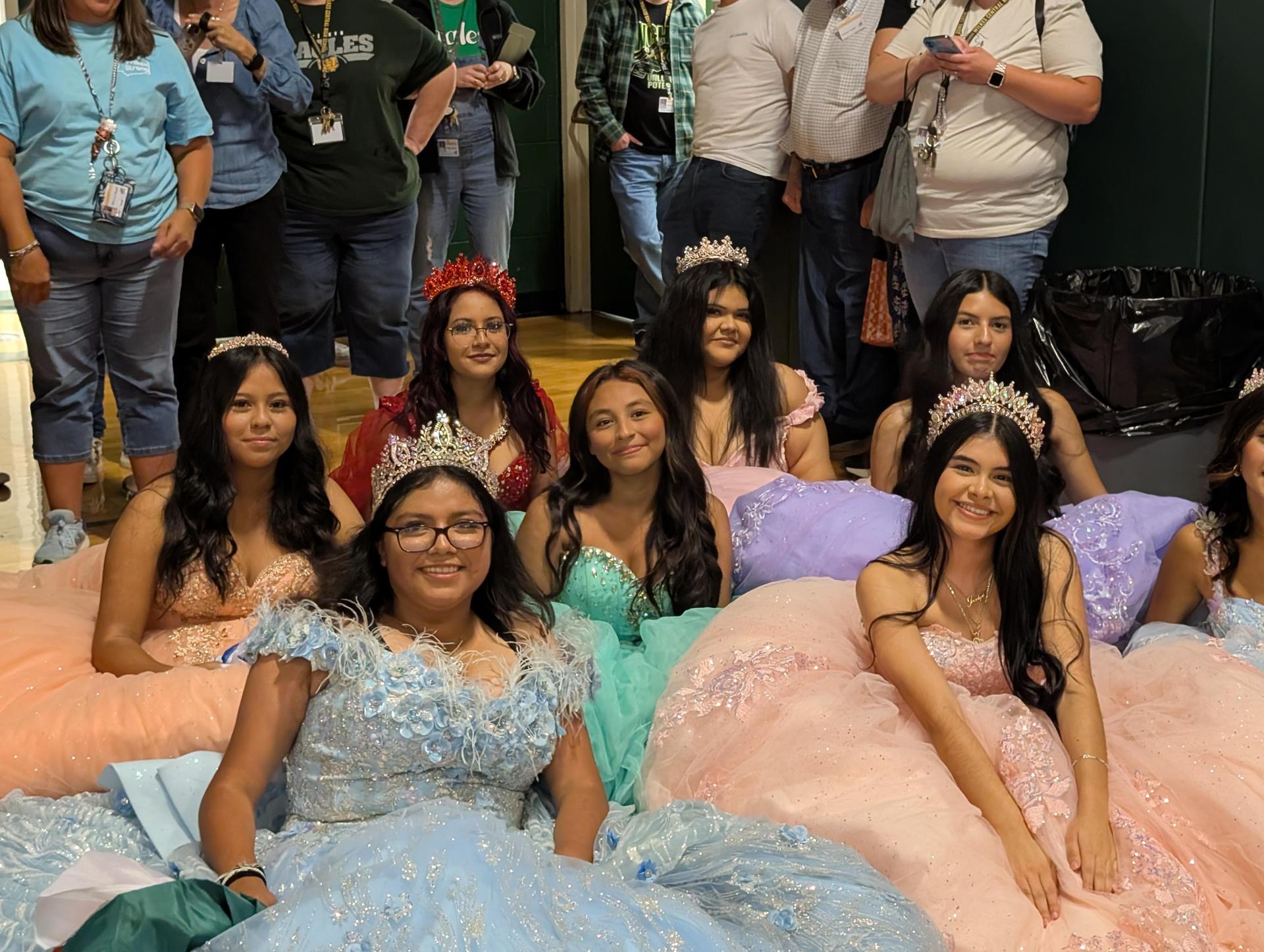 Group of girls wearing colorful gowns and crowns, sitting together at a school event.