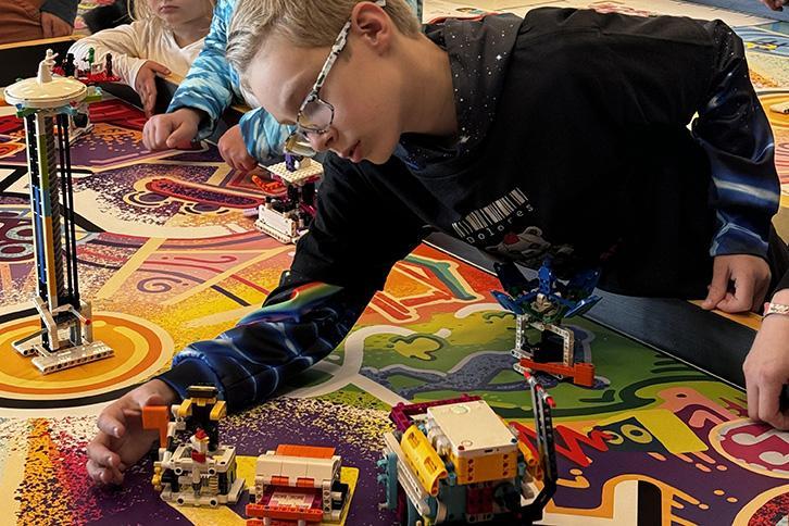 A student makes an adjustment to a LEGO build while onlookers stand around the large, colorful table.