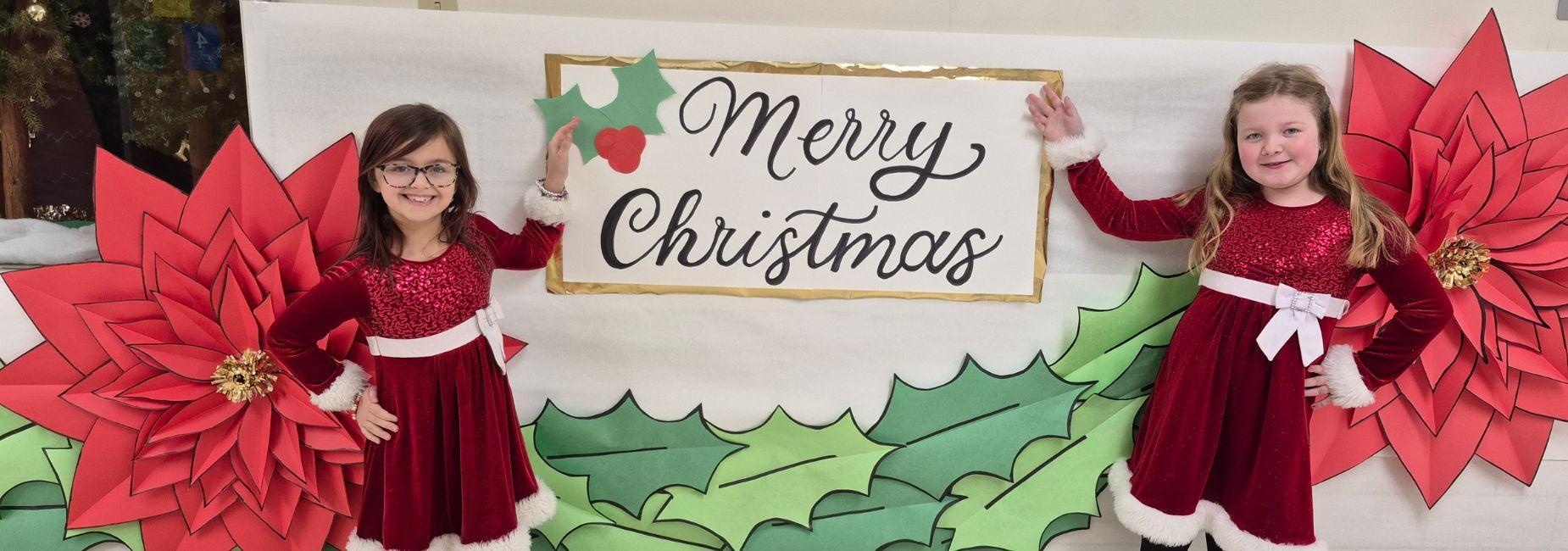Two girls in red dresses posing beside a Merry Christmas sign with holiday decorations.