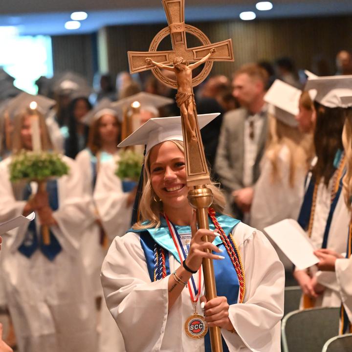 A smiling graduate in white robes holds a ceremonial staff during a graduation ceremony.