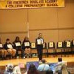 A speaker addressing an audience with students seated on stage holding signs.