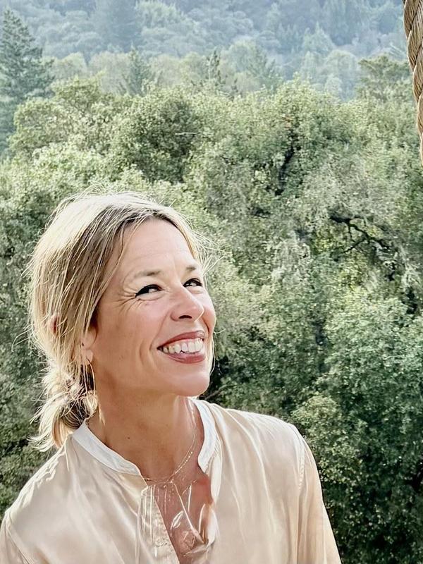 Smiling woman in a light blouse standing amidst lush greenery.