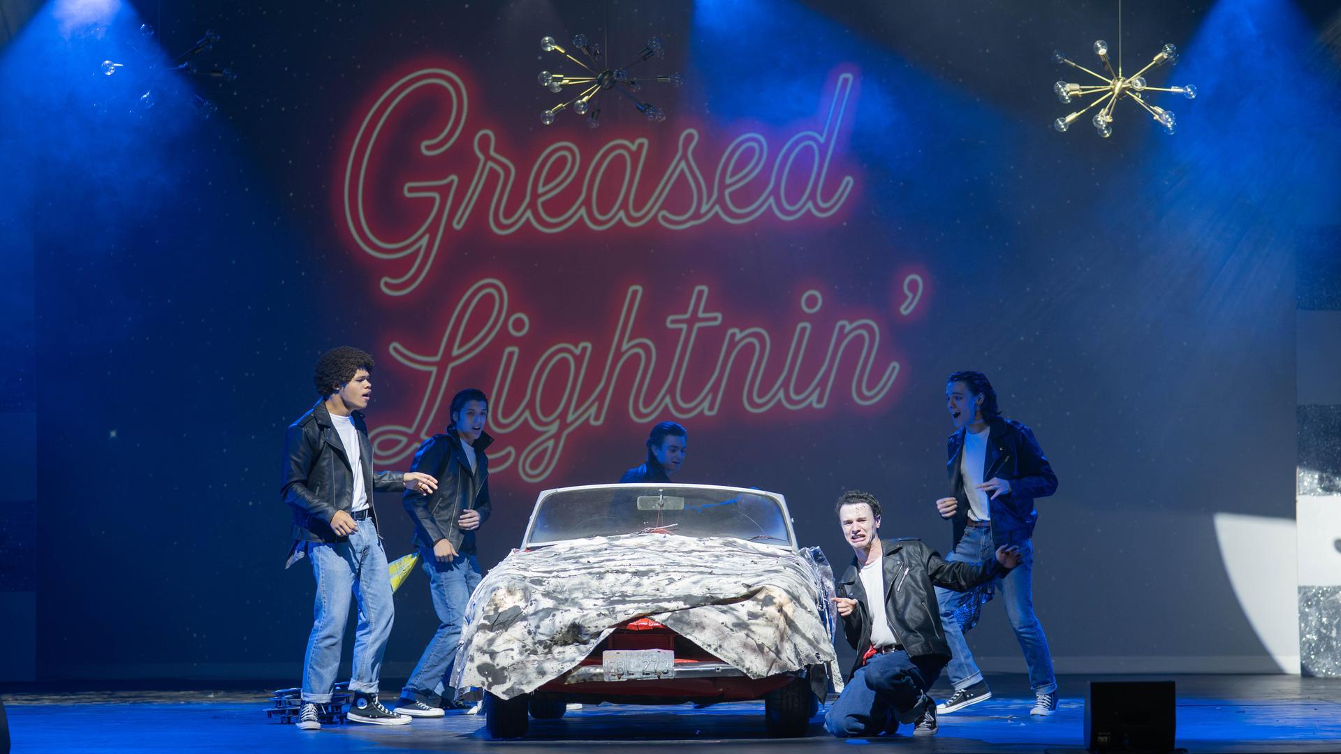 Musical scene with performers by a covered car and neon backdrop.