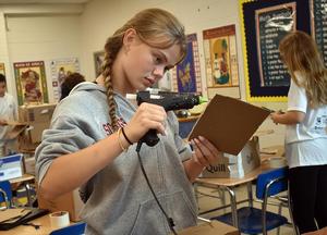 Girl using a glue gun on cardboard in a classroom.