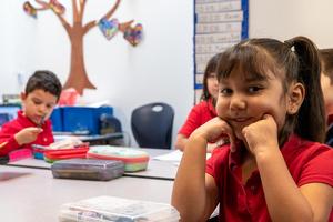 A student at Arco Iris Spanish Immersion School smiles for a photo. In the background, students work at desks in a classroom.
