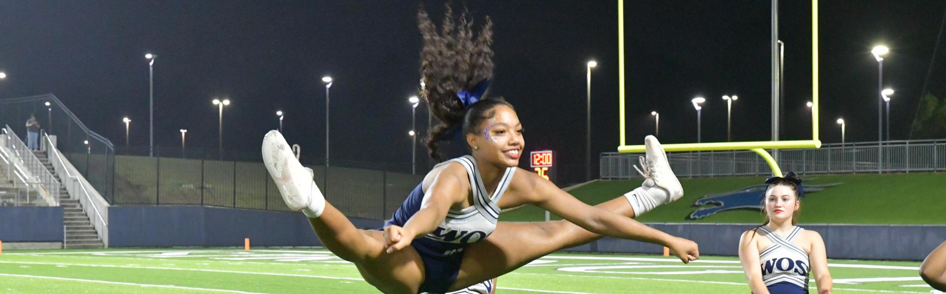 Cheerleader performing a high jump cheer with teammates in the background.