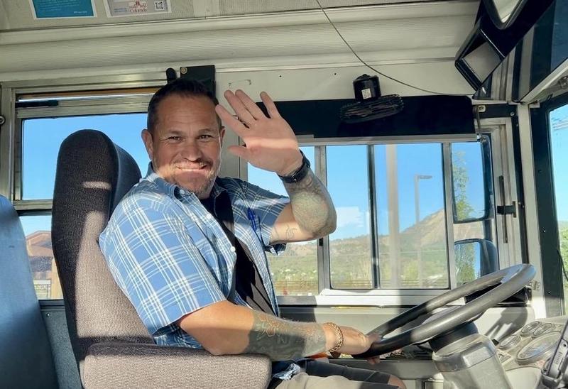 An adult waves from the driver's seat of a school bus