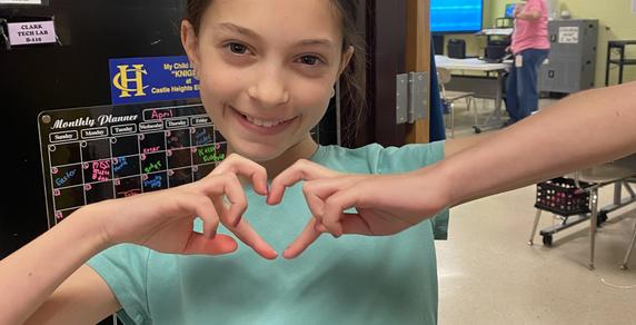 Girl in a classroom making a heart shape with her hands.