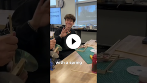 YouTube thumbnail of smiling student waving at the camera while building a mousetrap car in a classroom