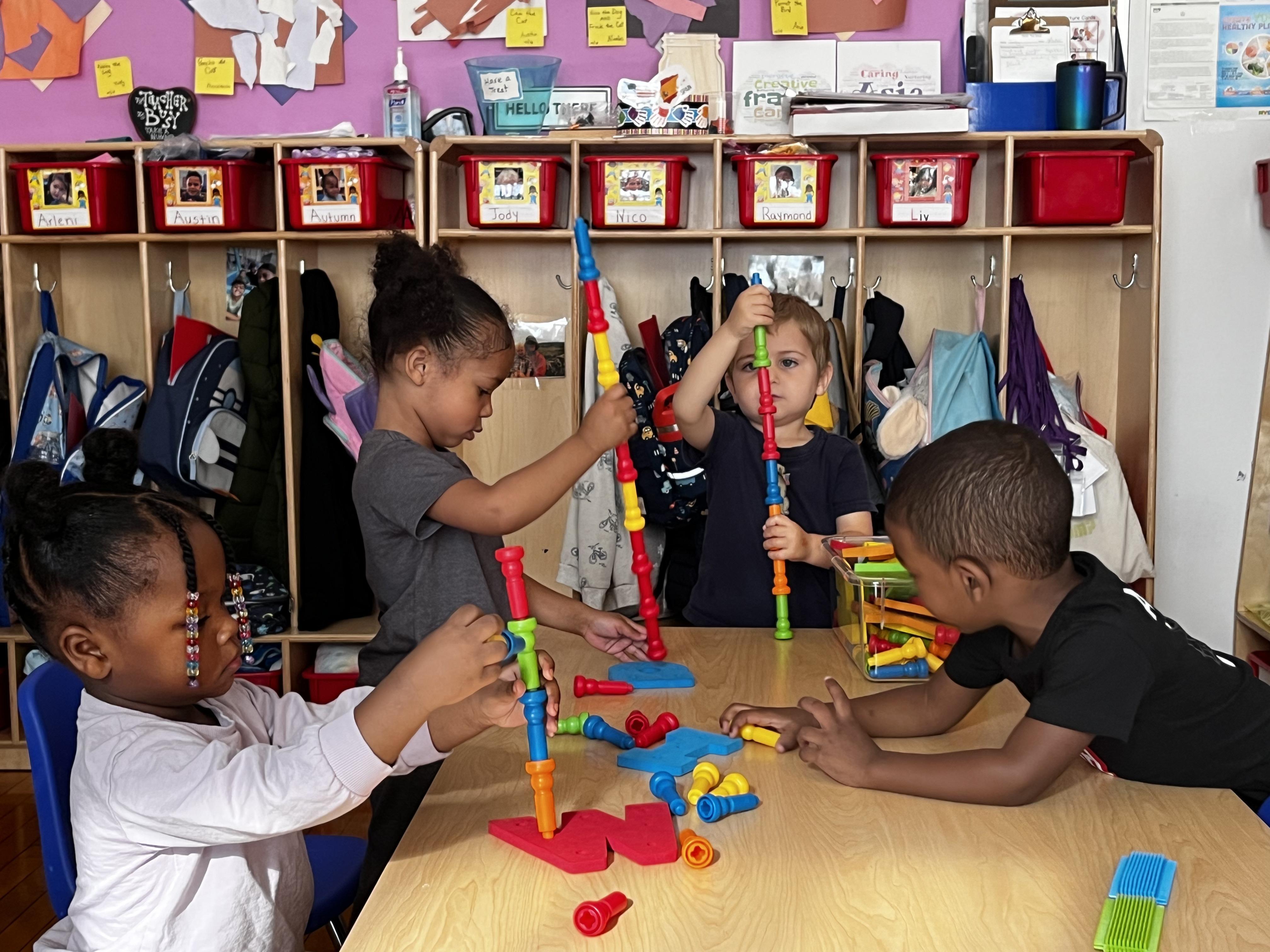 Four young children are seated at a table, focused on building vertical structures using colorful connecting blocks.