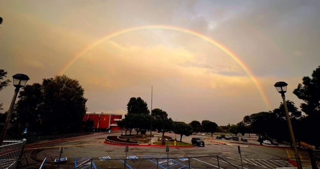A vibrant rainbow arcs over a cloudy sky with trees and a parking lot below.