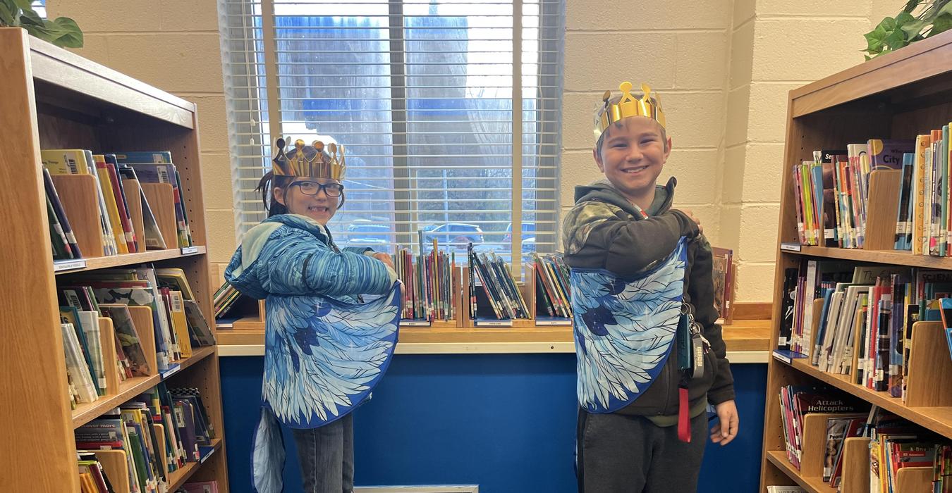 Two children in blue jay wing capes and crowns pose in a library aisle.