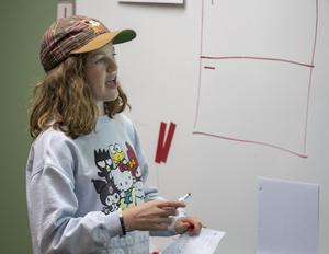 Student in hat at white board explaining math concepts.