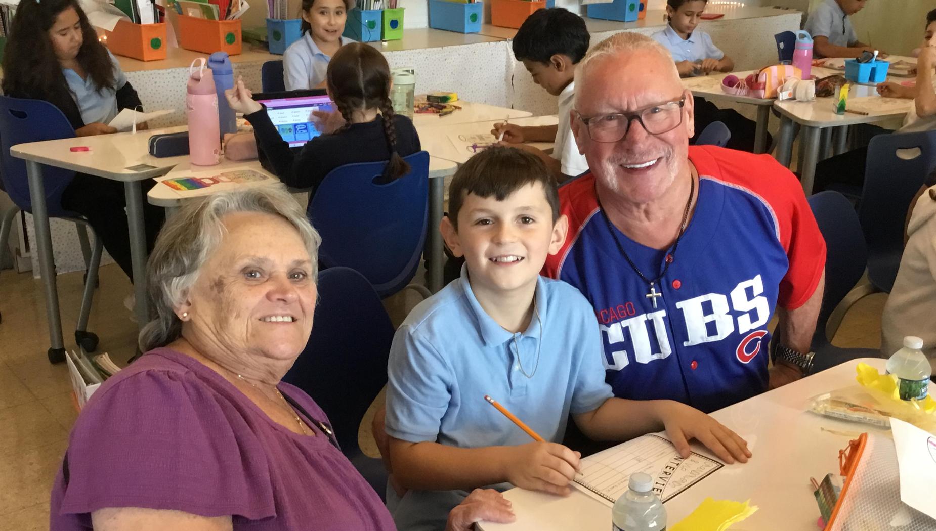 A smiling boy sits between two adults at a classroom table, engaged in an activity.