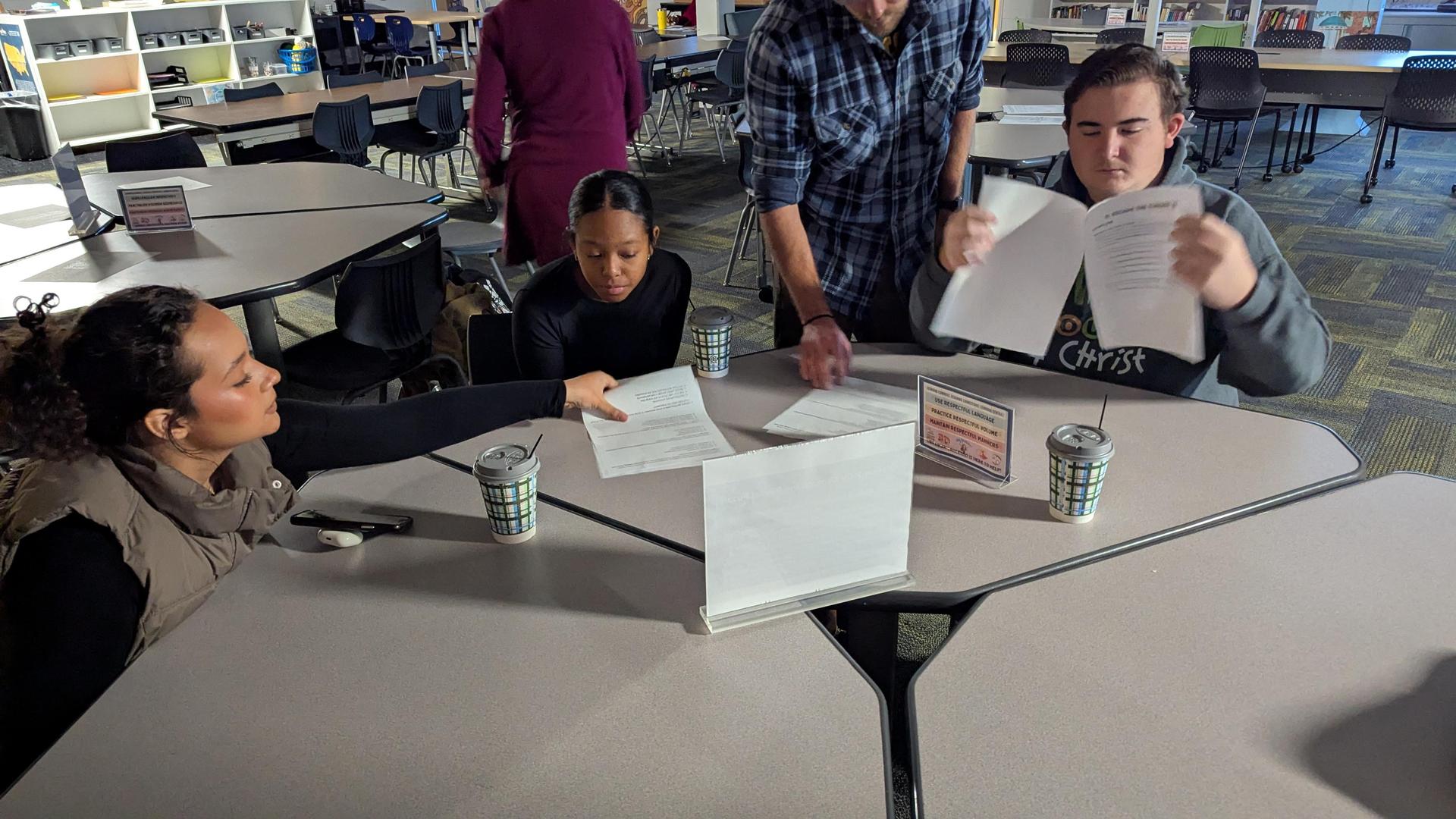Group of students collaborating at a table with papers and drinks.