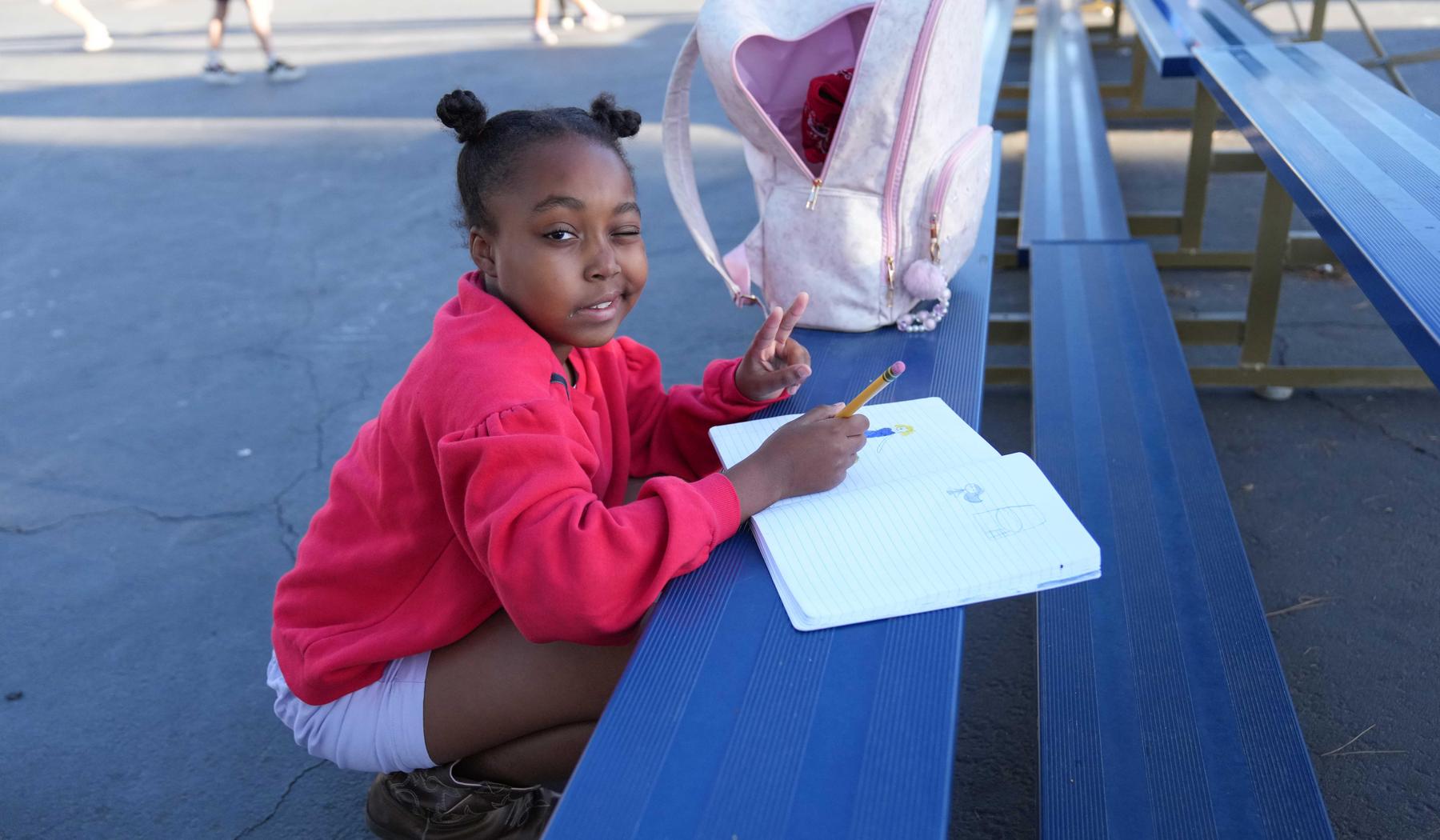 winking girl in red sweatshirt squatting while writing in workbook