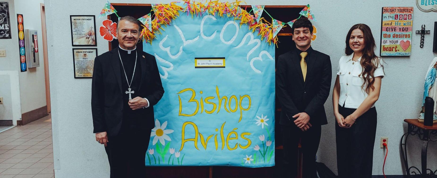 Three people stand beside a colorful sign welcoming Bishop Avil&eacute;s.