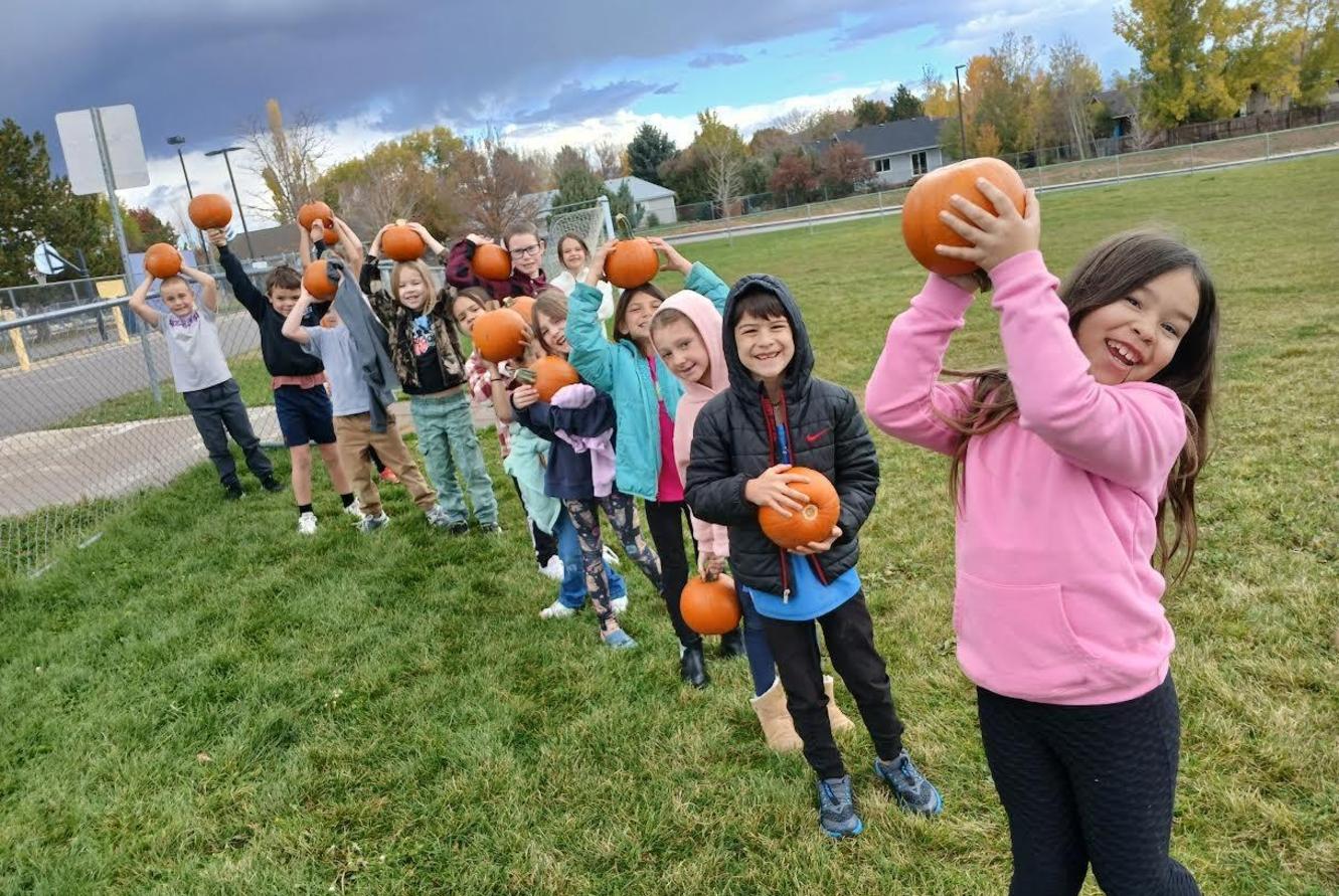Children lined up on a grassy field, each holding a pumpkin above their heads, smiling.