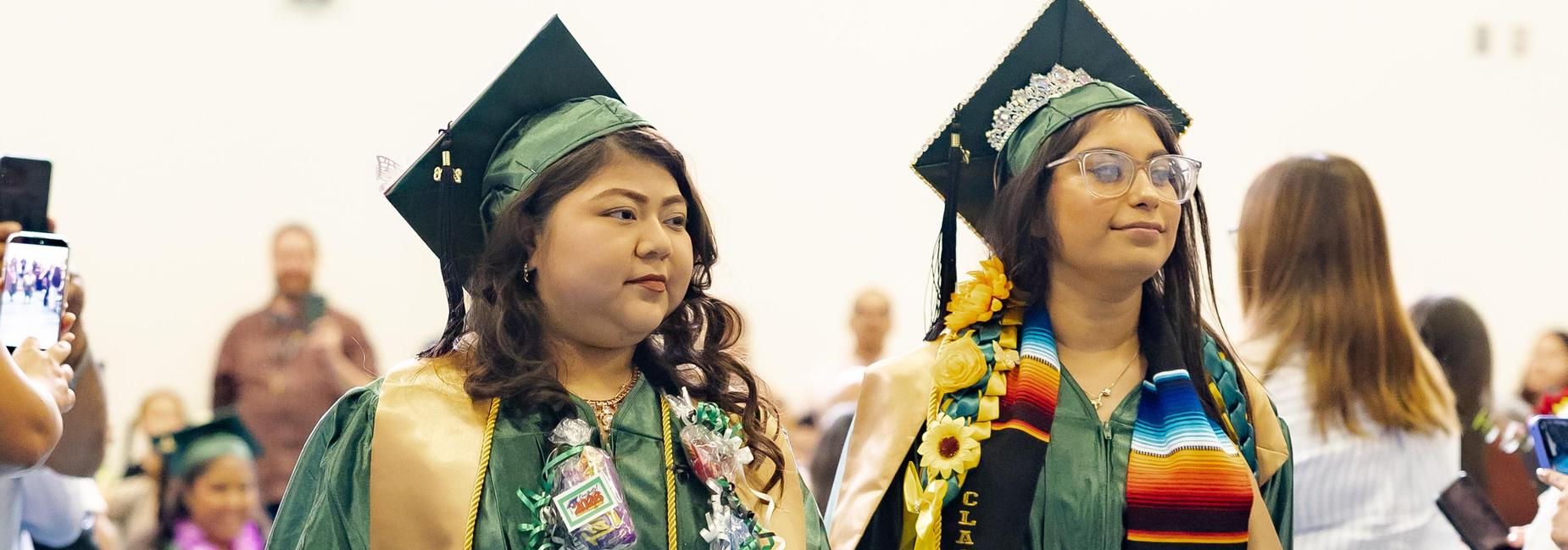 Two female graduates in green gowns walk hand-in-hand through the commencement processional. The graduate on the left wears a gold stole reading "Mi Cafecito." The one on the right wears a serape stole with sunflower accents and a small tiara on her cap. Both carry personal mementos and candy leis.