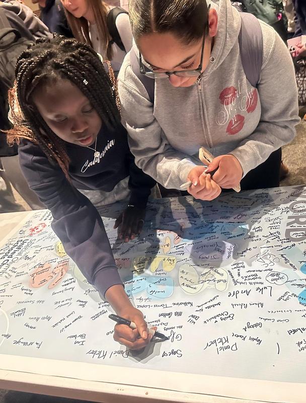 Two girls adding their signatures to a banner on a table