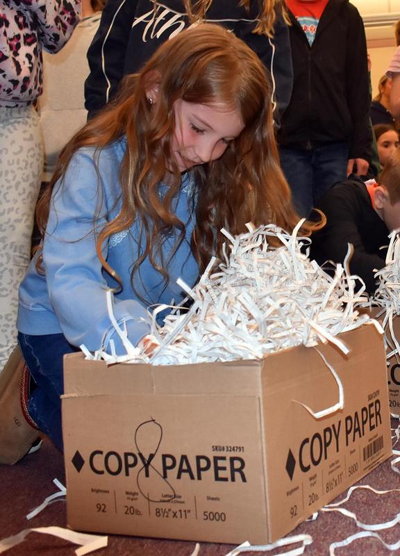 A girl with long hair examines a box filled with shredded paper on the floor.