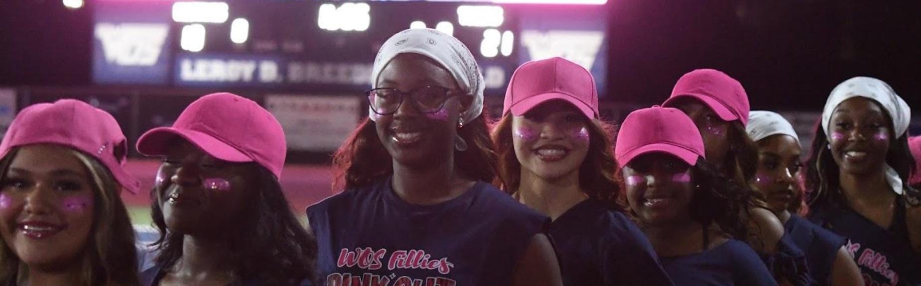 Cheerleaders in pink caps and outfits smiling while lining up at a football event.