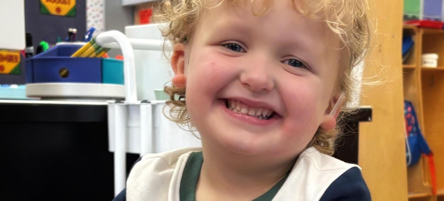 Smiling young boy with curly hair in a classroom setting.