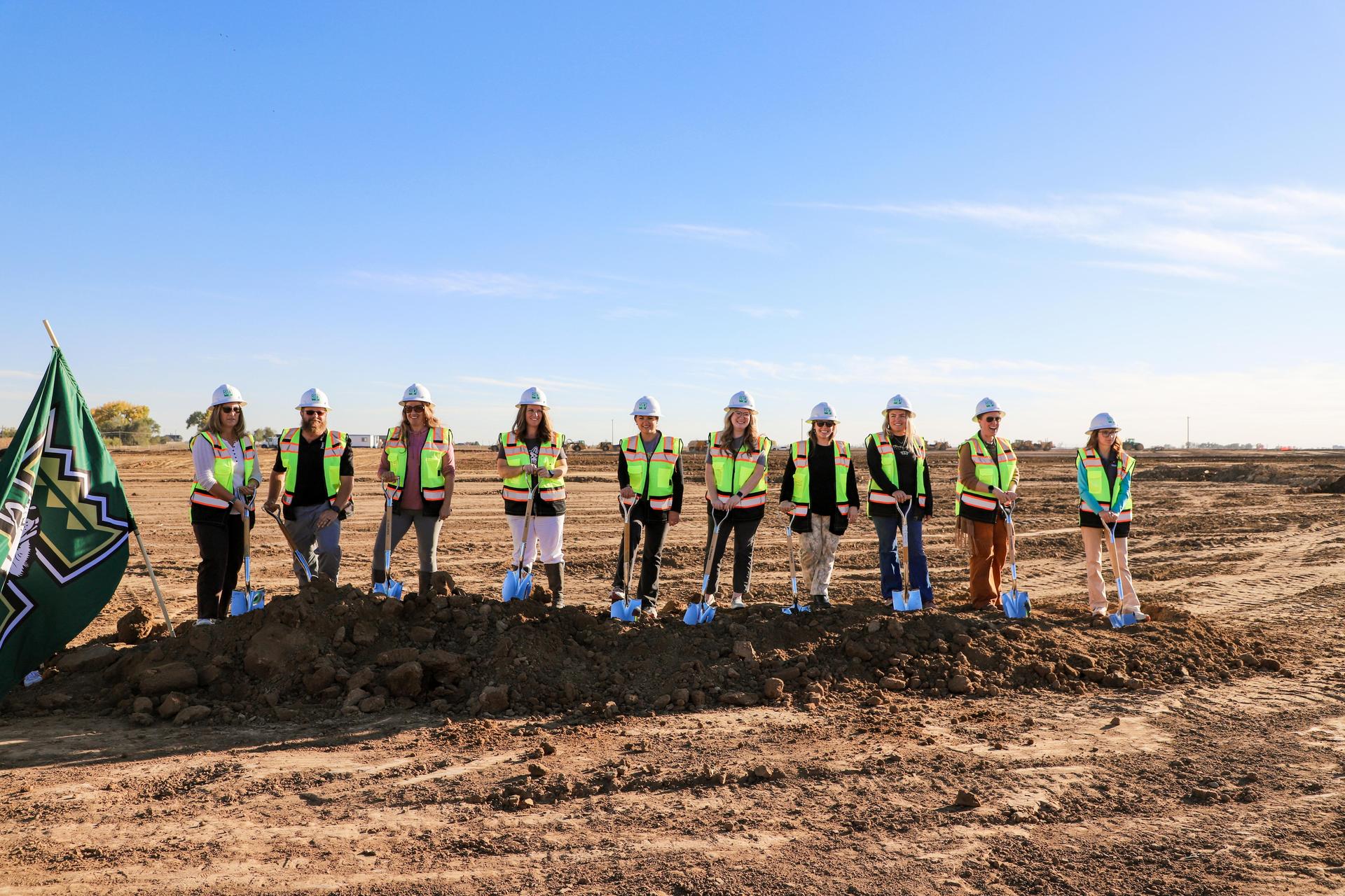 Groundbreaking ceremony with nine women in hard hats and reflective vests standing in a row.