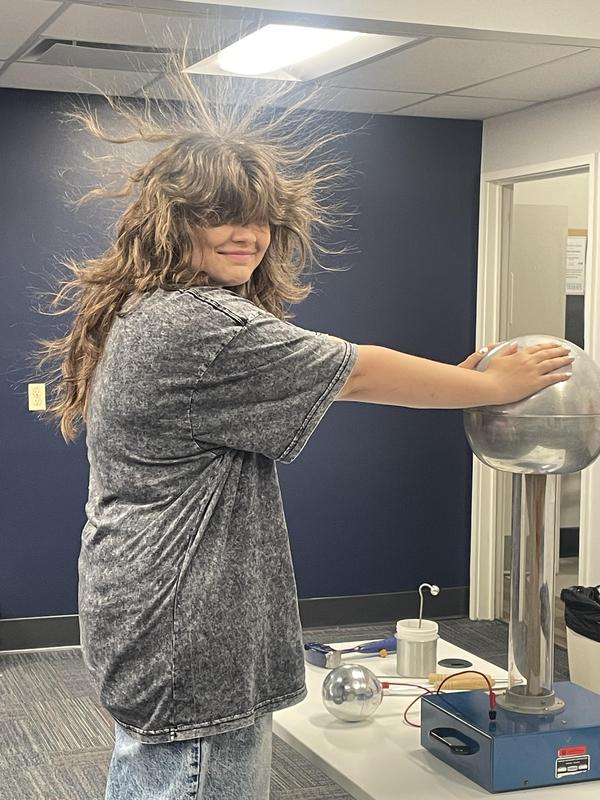 Kaydee has her hands on an elecronic generator with her hair standing on end.