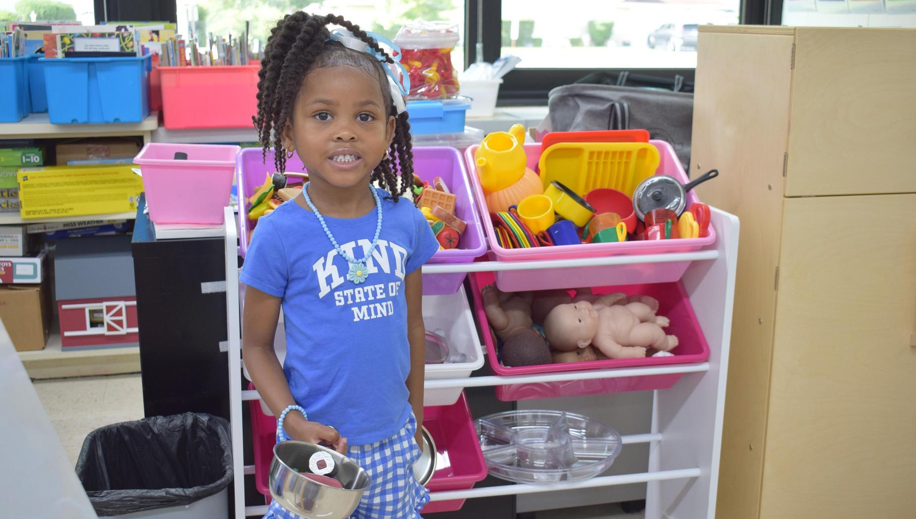 A young girl holding a pot in front of a play kitchen filled with toys.