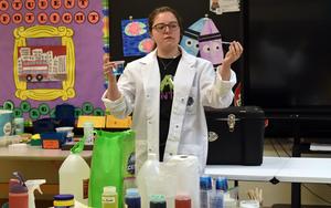 A woman in a lab coat holds a container while explaining to an audience in a classroom.