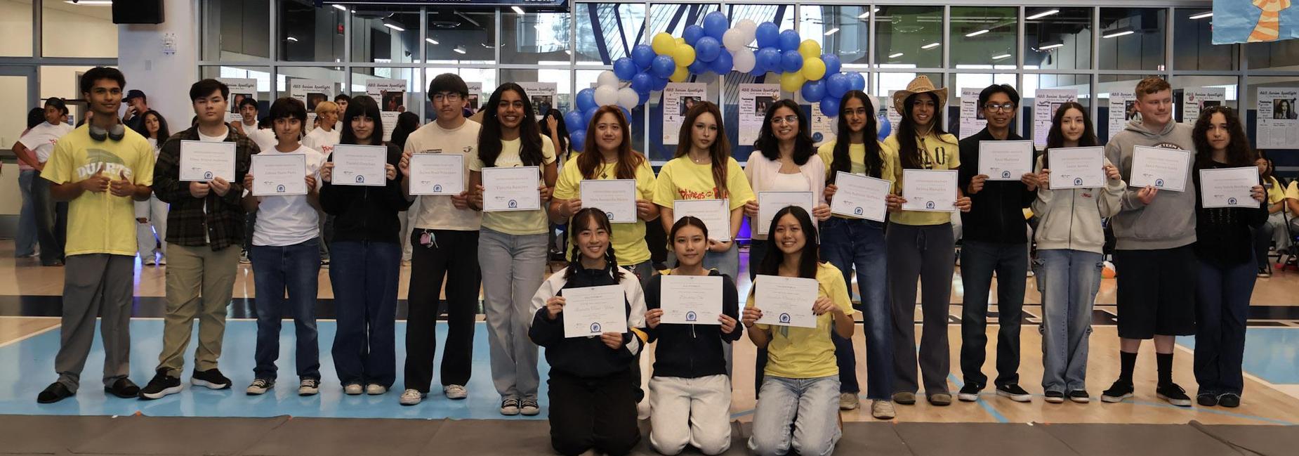 Group of students holding certificates in a gym decorated with balloons.