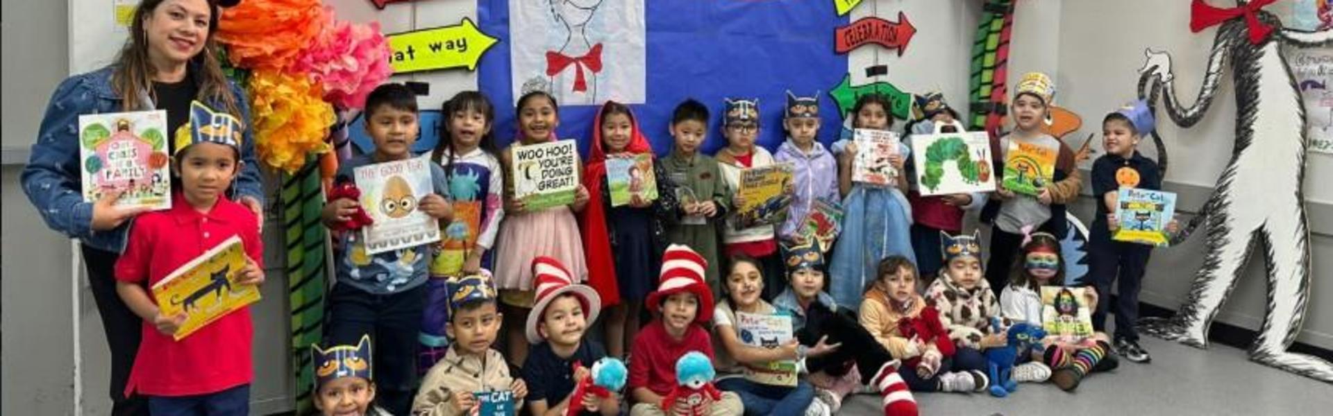 Group of children and an adult dressed in costumes, holding books in a colorful classroom.