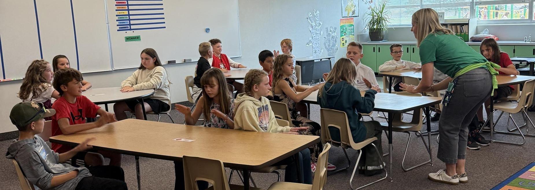 Children engaged in a classroom activity at tables, with a teacher leading.
