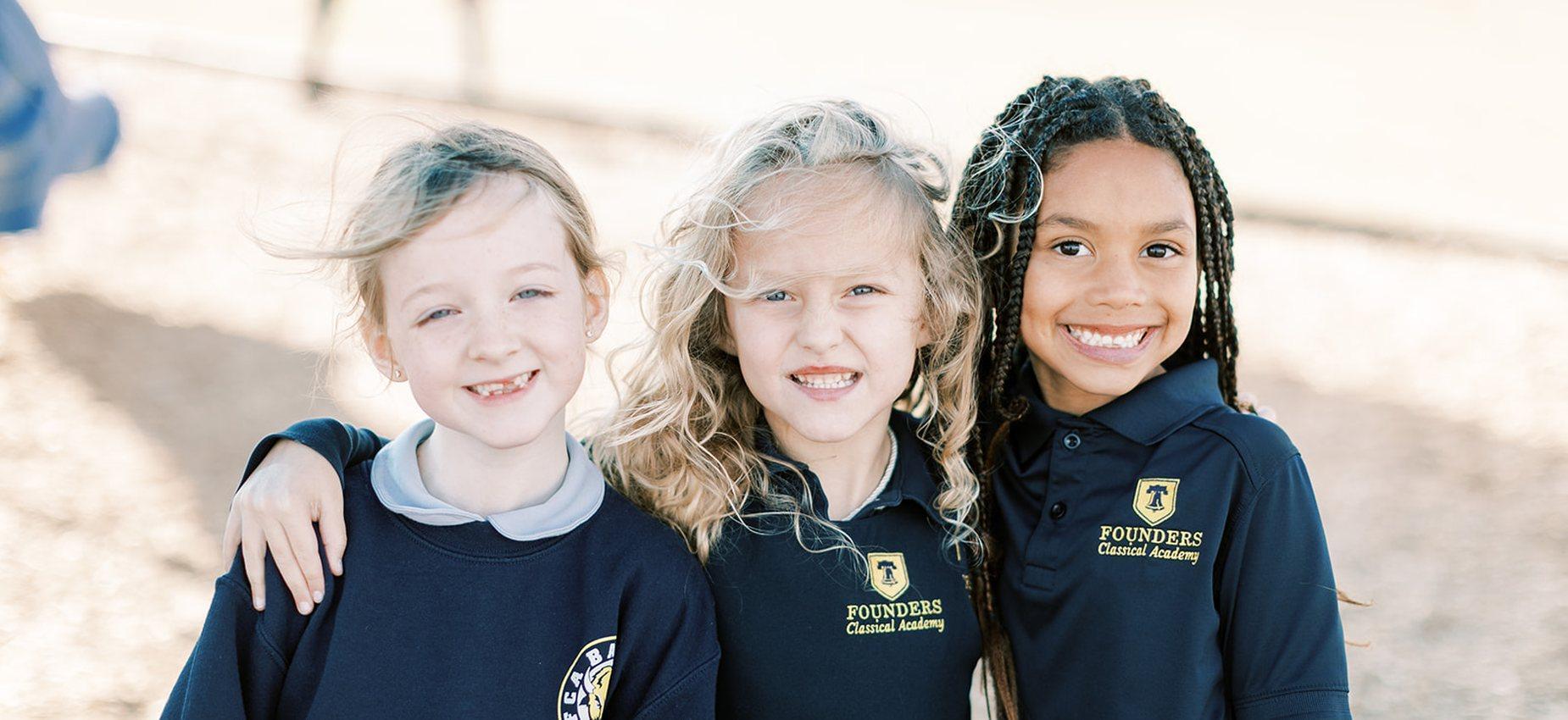 Three smiling elementary students in Founders uniforms stand arm in arm outside at Plainview Classical Academy