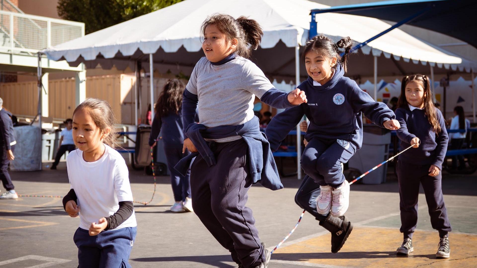 Two girls jumping rope excitedly, with others playing in the background.
