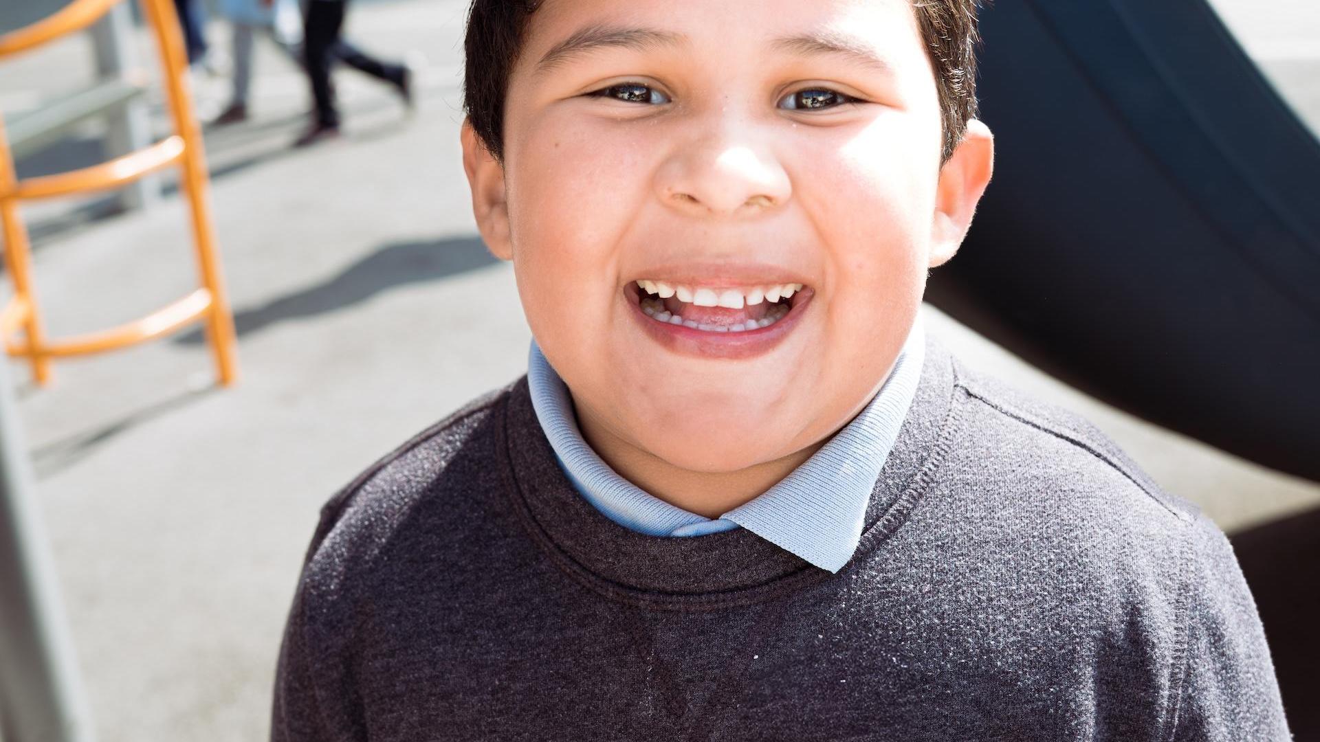 Smiling boy in a gray sweater, outdoors at a playground.