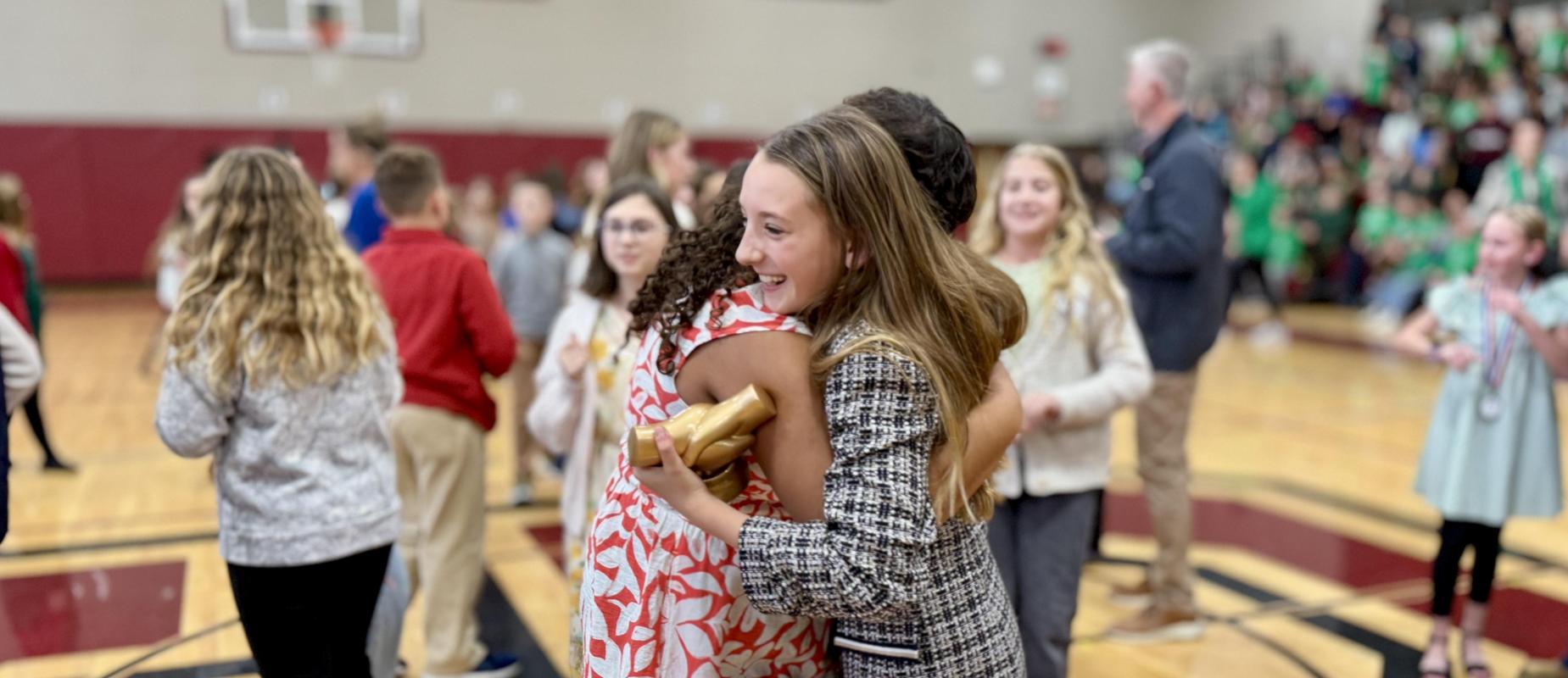 two girls smile and hug in gym with one holding trophy in her suit jacket, other in a floral dress