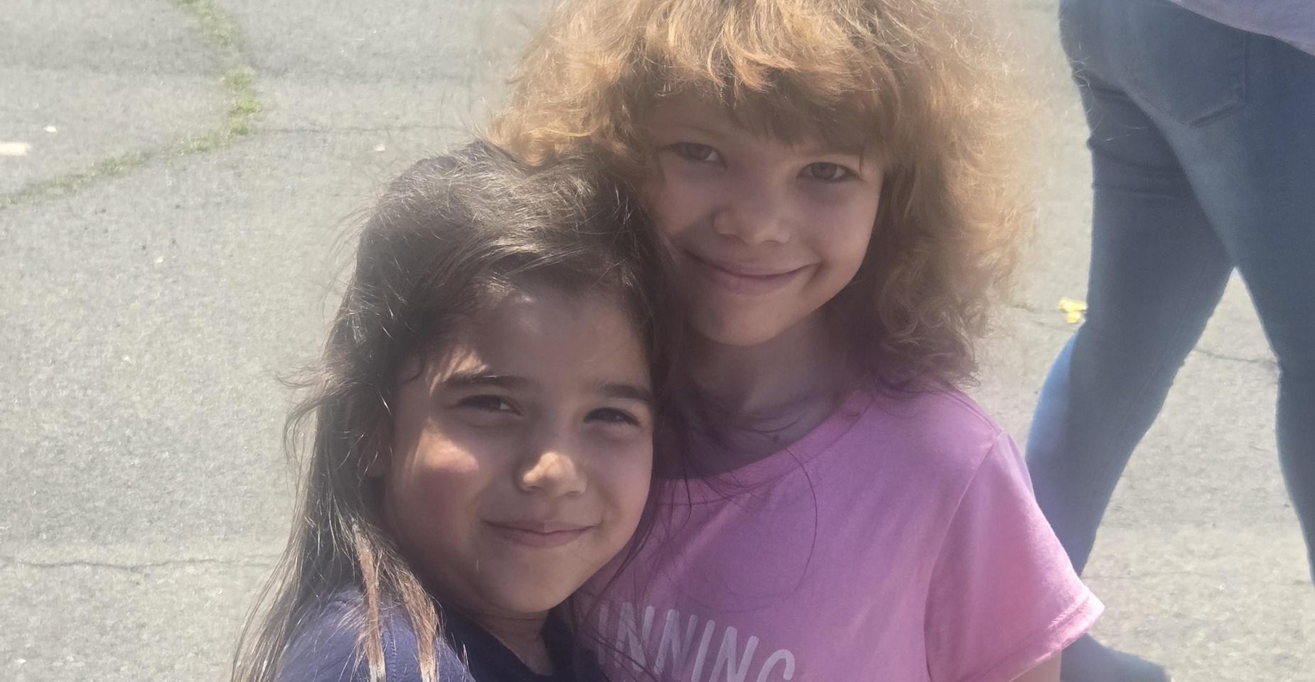 Two young girls smiling and hugging outdoors.