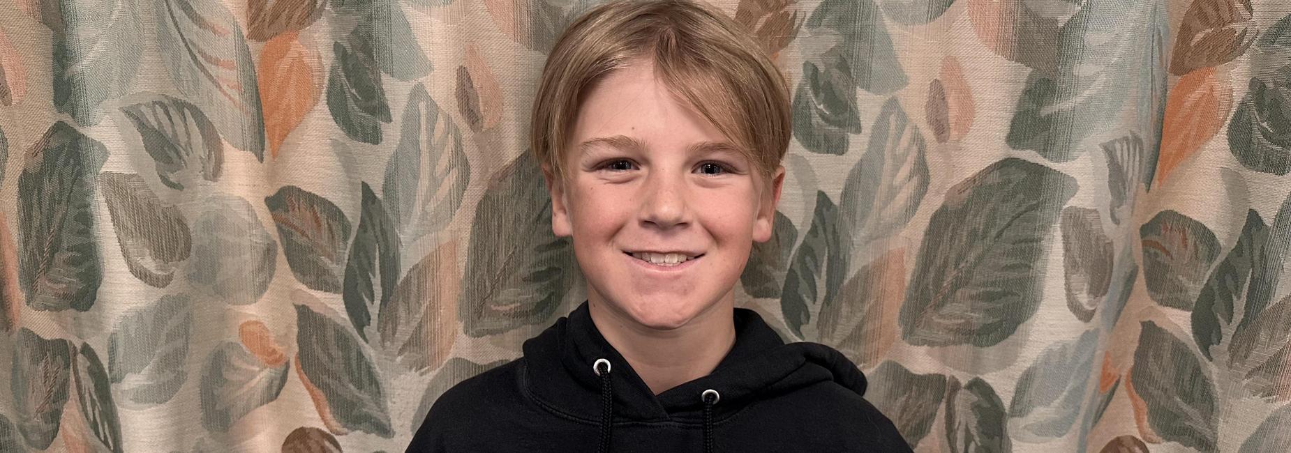 Smiling boy holding a certificate in front of a leafy curtain.