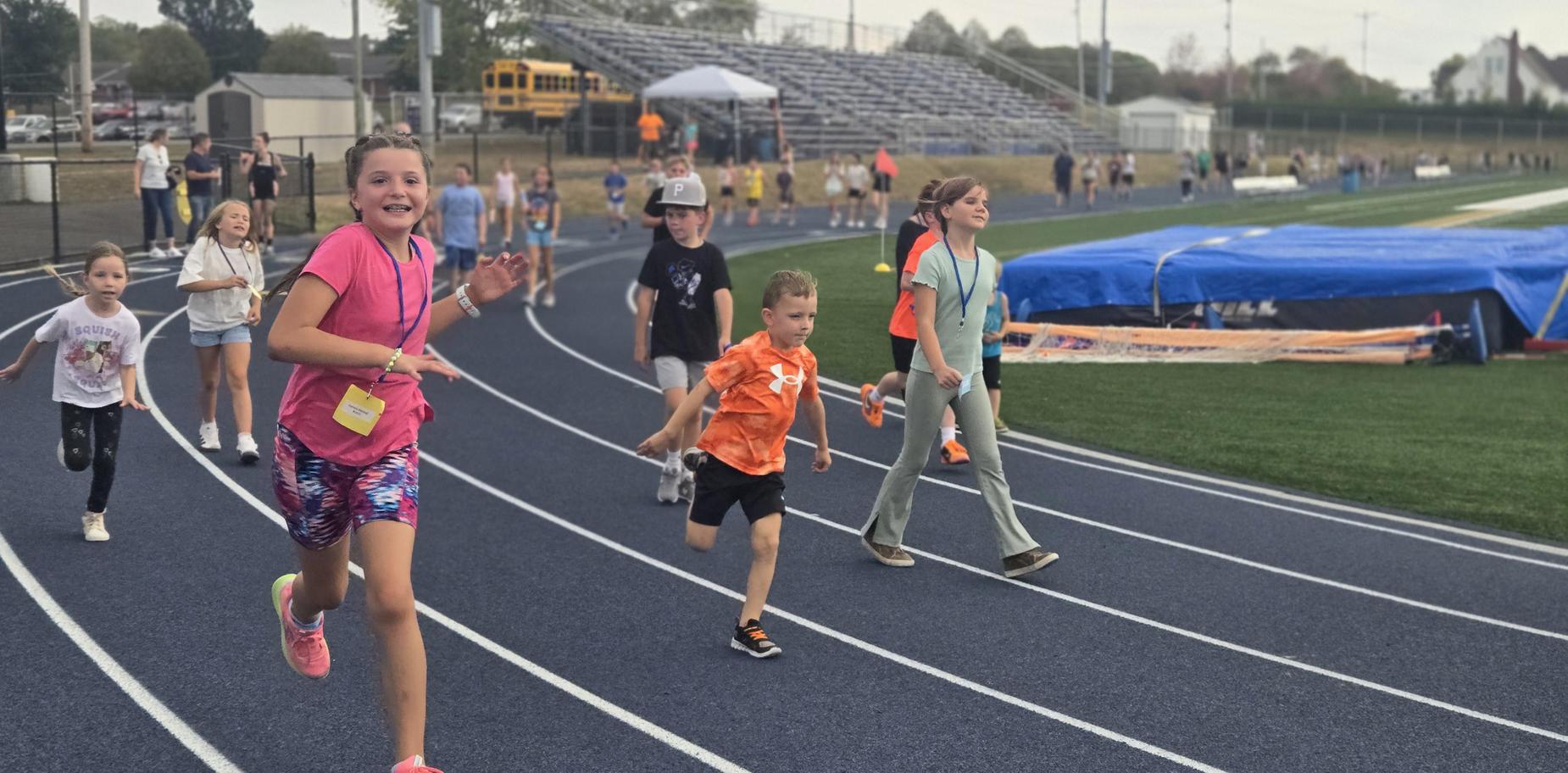 Kids run joyfully on the track, with a sports field and spectators nearby.